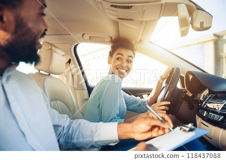 A driving instructor sits in the passenger seat of a car while a student driver sits in the drivers seat and turns to look at the instructor. The instructor is using a pen to write on a clipboard. 118437088