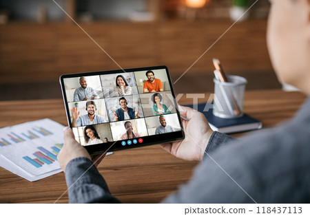 A person is sitting at a desk and looking at a tablet computer. The tablet is displaying a video conference with eight people. The people are all smiling and looking at the camera. 118437113