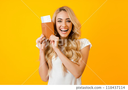 A young woman with long blonde hair stands, against a bright yellow background and smiles excitedly as she holds a passport and boarding pass in her hands. A young woman with long blonde hair stands, against a bright yellow background and smiles excitedly as she holds a passport and boarding pass in her hands. 118437114