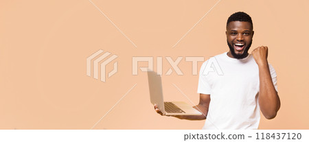 African American man holding a laptop computer in front of a peach-colored background. He appears to be celebrating a success or achievement, panorama with copy space 118437120