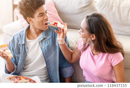 A teen couple is sitting on the floor in a living room, sharing a pizza. The girl is feeding a slice to the guy, and they both look happy and relaxed, closeup 118437155