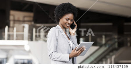 Attractive concentrated african businesswoman making phone call and looking at digital tablet screen while standing near modern office building. 118437169