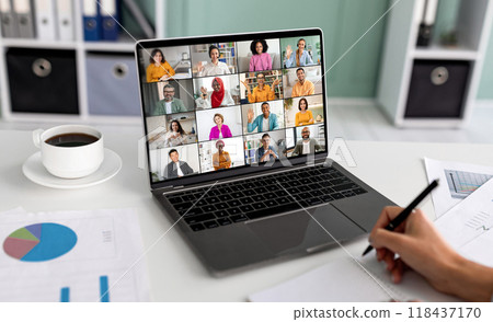 A laptop screen displays a video conference meeting with a grid of multiple participants, all of whom are smiling and waving. Computer is positioned on a white desk in front of a person who is writing 118437170