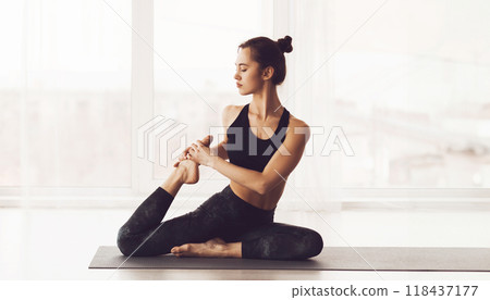 A young woman is depicted sitting on a yoga mat in a well-lit studio. She is dressed in black athletic wear and is performing a yoga pose, maintaining a focused yet serene expression 118437177