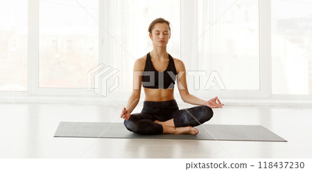 A woman in athletic clothing sits cross-legged on a gray yoga mat, meditating in a bright, sunlit room with large windows. A woman in athletic clothing sits cross-legged on a gray yoga mat, meditating in a bright, sunlit room with large windows. 118437230