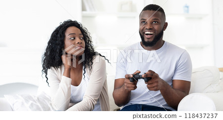 A couple sits on a couch, the man is engrossed in playing a video game, while the woman watches with a look of annoyance. 118437237