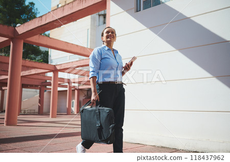 Confident businesswoman walking outdoors with a laptop bag and smartphone in a modern office park 118437962