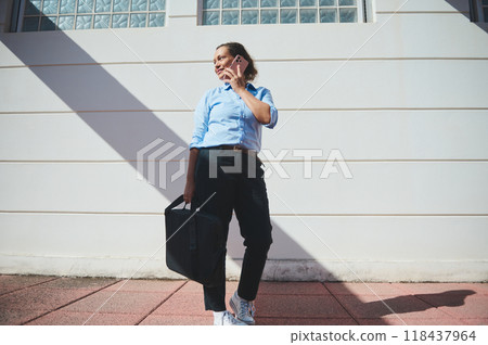 Confident professional woman standing outdoors, talking on smartphone, carrying a business bag, wearing casual attire. Strong shadows from the afternoon sun create a dynamic visual effect. Confident professional woman standing outdoors, talking on smartphone, carrying a business bag, wearing casual attire. Strong shadows from the afternoon sun create a dynamic visual effect. 118437964
