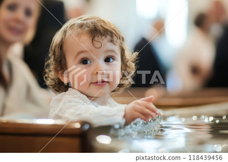 A wide-eyed toddler with curly hair in a white lace dress reaches towards sparkling baptismal water, surrounded by blurred figures of adults 118439456