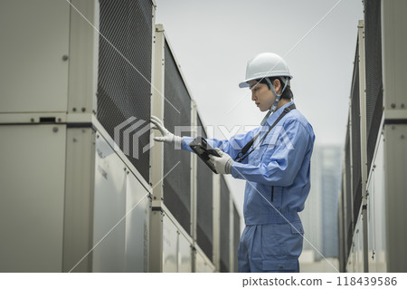 Building maintenance image: A man in work clothes inspecting the outdoor air conditioning unit installed on the roof of a building 118439586