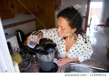Woman preparing a blend of fresh ingredients in a kitchen setting, pouring water into a blender for a homemade recipe creation 118440432
