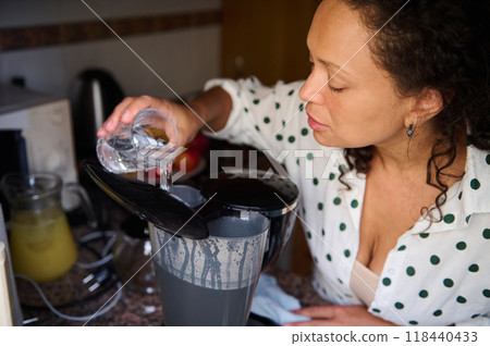 Woman preparing coffee by pouring water into a modern coffee maker in a cozy kitchen setting Woman preparing coffee by pouring water into a modern coffee maker in a cozy kitchen setting 118440433