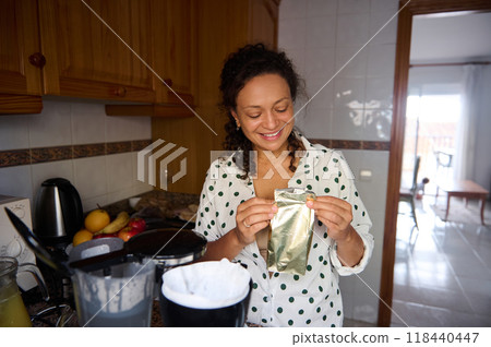 Smiling woman in patterned pajamas preparing a meal in her cozy kitchen, surrounded by fresh fruits and kitchen appliances Smiling woman in patterned pajamas preparing a meal in her cozy kitchen, surrounded by fresh fruits and kitchen appliances 118440447