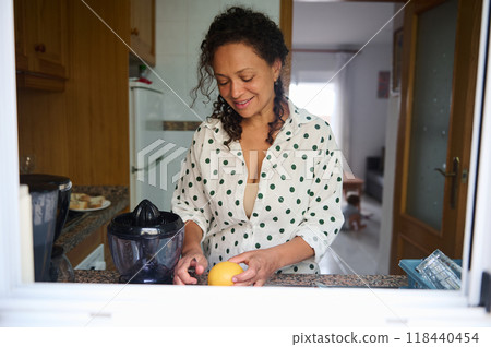 Woman preparing fresh citrus juice in cozy kitchen with sunny window, exuding warmth and homely atmosphere 118440454