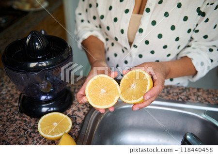 Woman preparing fresh orange juice in kitchen with electric juicer and halved oranges for a healthy drink. 118440458