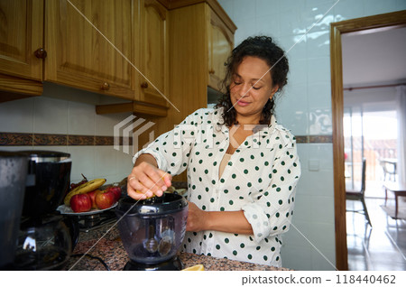Woman in polka dot blouse using a blender in a cozy kitchen with fresh fruits on the counter, enjoying a relaxed morning routine 118440462