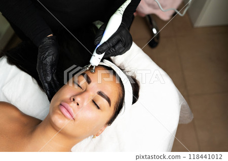Close-up portrait of a beautiful woman with closed eyes and apparatus for skin rejuvenation. Beauty salon procedures. RF lifting. Microcurrents. 118441012