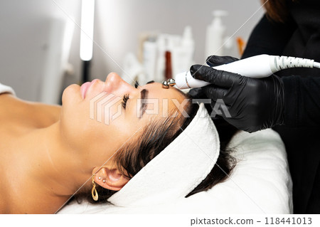 Close-up portrait of a beautiful woman with closed eyes and apparatus for skin rejuvenation. Beauty salon procedures. RF lifting. Microcurrents. 118441013
