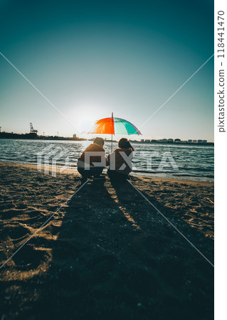 A boy and a girl sharing a colorful umbrella under the sunlight of the seaside sunset A boy and a girl sharing a colorful umbrella under the sunlight of the seaside sunset 118441470