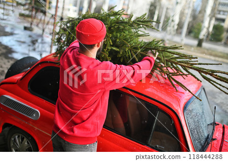a young man in a red sweater and a red hat puts a Christmas tree on the roof of a red retro car against the background of falling snow. a young man in a red sweater and a red hat puts a Christmas tree on the roof of a red retro car against the background of falling snow. 118444288