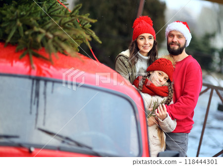 family - mother, daughter and father in Santa's hat are hugging and smiling on the background of a red retro car with a Christmas tree on the roof. family - mother, daughter and father in Santa's hat are hugging and smiling on the background of a red retro car with a Christmas tree on the roof. 118444301
