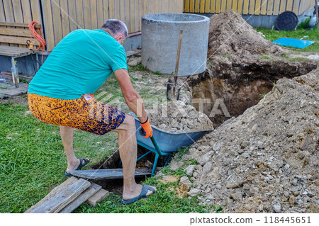 When digging pit for septic tank made of concrete rings, homeowner uses bayonet shovel and garden wheelbarrow. 118445651