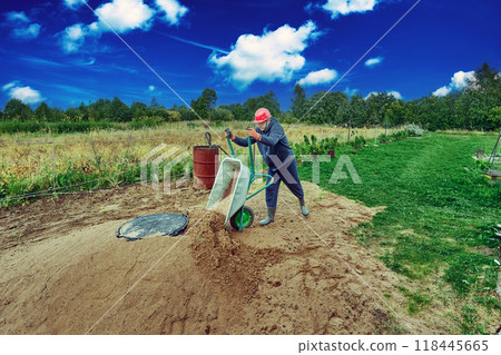 Worker pours sand from wheelbarrow onto concrete ring of septic tank. 118445665