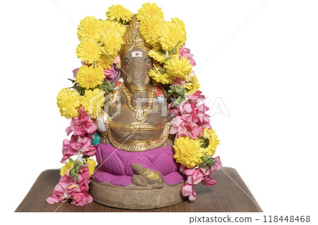Hindu god Ganesha idol made of clay on wooden table on white background decorated with flower garlands at home in celebration of Ganesha Chaturthi festival. Closeup Hindu god Ganesha idol made of clay on wooden table on white background decorated with flower garlands at home in celebration of Ganesha Chaturthi festival. Closeup 118448468
