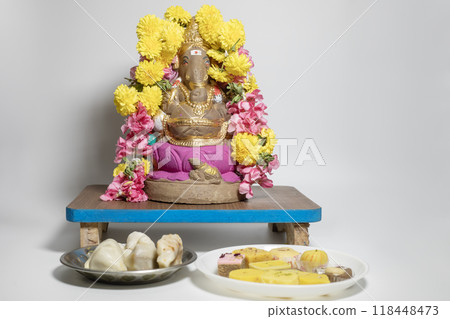Ganesha idol made of clay decorated with flower garlands on wooden table with sweets on white background. Closeup Ganesha idol made of clay decorated with flower garlands on wooden table with sweets on white background. Closeup 118448473
