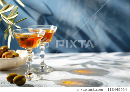 Elegant Cocktails and Olives. Two glasses of amber liquid and a bowl of olives on a white surface against a blue backdrop with shadows. 118449934
