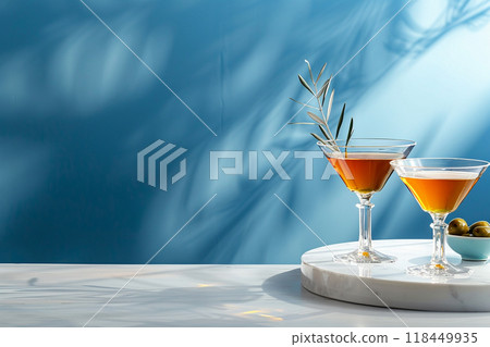 Elegant Martini Glasses. Two glasses of amber liquid and a bowl of olives on a white surface against a blue backdrop with shadows. Elegant Martini Glasses. Two glasses of amber liquid and a bowl of olives on a white surface against a blue backdrop with shadows. 118449935