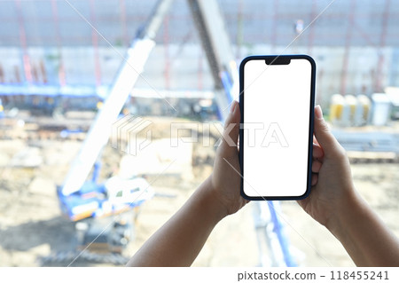 Close up of hands holding smartphone with white empty screen on blurred construction site background Close up of hands holding smartphone with white empty screen on blurred construction site background 118455241