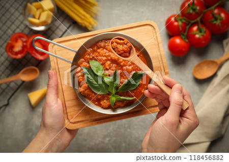 Bolognese sauce in pan and spoon in hands, ingredients on gray background, top view Bolognese sauce in pan and spoon in hands, ingredients on gray background, top view 118456882