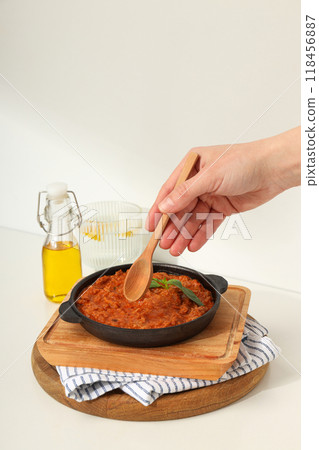 Bolognese sauce in frying pan, wooden boards, bottle of oil and hand with spoon on light background Bolognese sauce in frying pan, wooden boards, bottle of oil and hand with spoon on light background 118456887