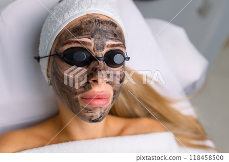 Close-up of cosmetologist's hand making hardware carbon peeling by young pretty customer woman in safety glasses headdress lying on couch in beauty salon. Facial cleaning concept. Cosmetology Services 118458500