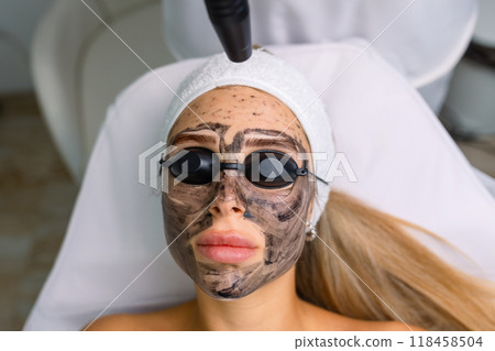 Close-up of cosmetologist's hand making hardware carbon peeling by young pretty customer woman in safety glasses headdress lying on couch in beauty salon. Facial cleaning concept. Cosmetology Services Close-up of cosmetologist's hand making hardware carbon peeling by young pretty customer woman in safety glasses headdress lying on couch in beauty salon. Facial cleaning concept. Cosmetology Services 118458504
