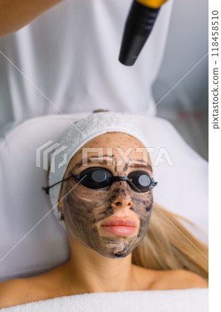 Close-up of cosmetologist's hand making hardware carbon peeling by young pretty customer woman in safety glasses headdress lying on couch in beauty salon. Facial cleaning concept. Cosmetology Services Close-up of cosmetologist's hand making hardware carbon peeling by young pretty customer woman in safety glasses headdress lying on couch in beauty salon. Facial cleaning concept. Cosmetology Services 118458510