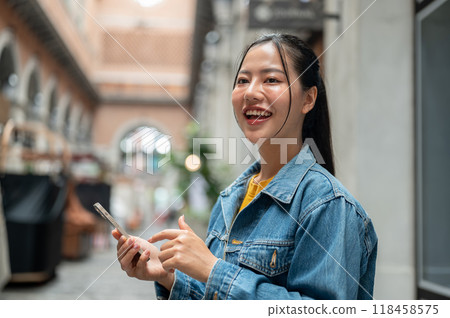 A beautiful Asian woman stands outdoors, holding a smartphone and smiling while looking away. 118458575