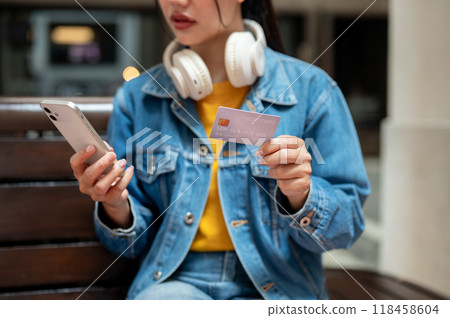A cropped image of a woman sitting on a bench in the city, holding a credit card and smartphone. A cropped image of a woman sitting on a bench in the city, holding a credit card and smartphone. 118458604