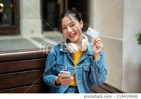 A beautiful Asian woman sits on a bench outdoors, holding up a credit card and smiling at the camera A beautiful Asian woman sits on a bench outdoors, holding up a credit card and smiling at the camera 118458606