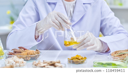 Scientist carefully holds and observes flask filled with yellow liquid, surrounded on the table by herbal ingredients contained in transparent petri dishes. Sample image for experiments in lap setting 118459411