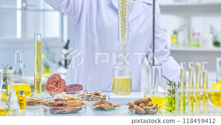 A scientist wearing a lab coat, gloves and a protective mask is holding a flask filled with a yellow liquid, examining it in a lab surrounded by herbs, flasks and other lab equipment. 118459412