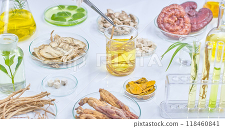 Sample photo of an experimental setting with scientist picking a strand of herbs from a petri dish containing yellow solution placed on a table next to other herbs contained in experimental equipment. 118460841