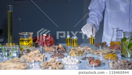 Modern lab setting with gray background features a scientist carefully picks up a vial containing a yellow liquid. The lab table is filled with various herbs, roots, and lab equipment. 118460844