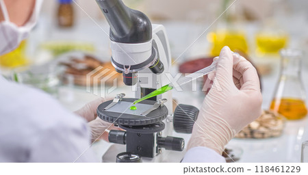 Rear view of a scientist dropping green extract onto a microscope for observation. Laboratory setting with herbs and instruments on the table, suitable for experimental photography. 118461229