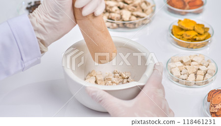 A scientist hand with a wooden pestle grinds herbal pieces in a white mortar. Surrounding ingredients showcase a vibrant array of natural herbs in petri dish. 118461231