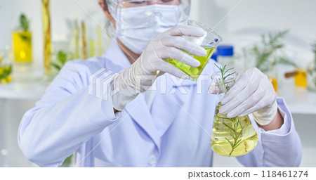 A scientist carefully pours a green liquid from a beaker into a flask containing a rosemary sprig and glass containers containing herbs in the backdrop. The picture show experiments in a lab setting. 118461274