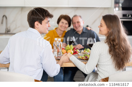 Couple sharing close moment at table, holding hands and locking eyes, while happy parents watching in background at family home. Blessing of parents Couple sharing close moment at table, holding hands and locking eyes, while happy parents watching in background at family home. Blessing of parents 118461817