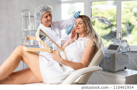 Young woman looking in mirror while experienced cosmetologist explaining facial procedure 118461954