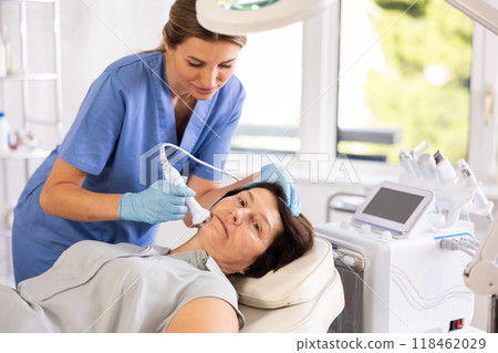 Cosmetologist performing anti-aging face hardware therapy procedure for aged female client in clinic of aesthetic medicine Cosmetologist performing anti-aging face hardware therapy procedure for aged female client in clinic of aesthetic medicine 118462029